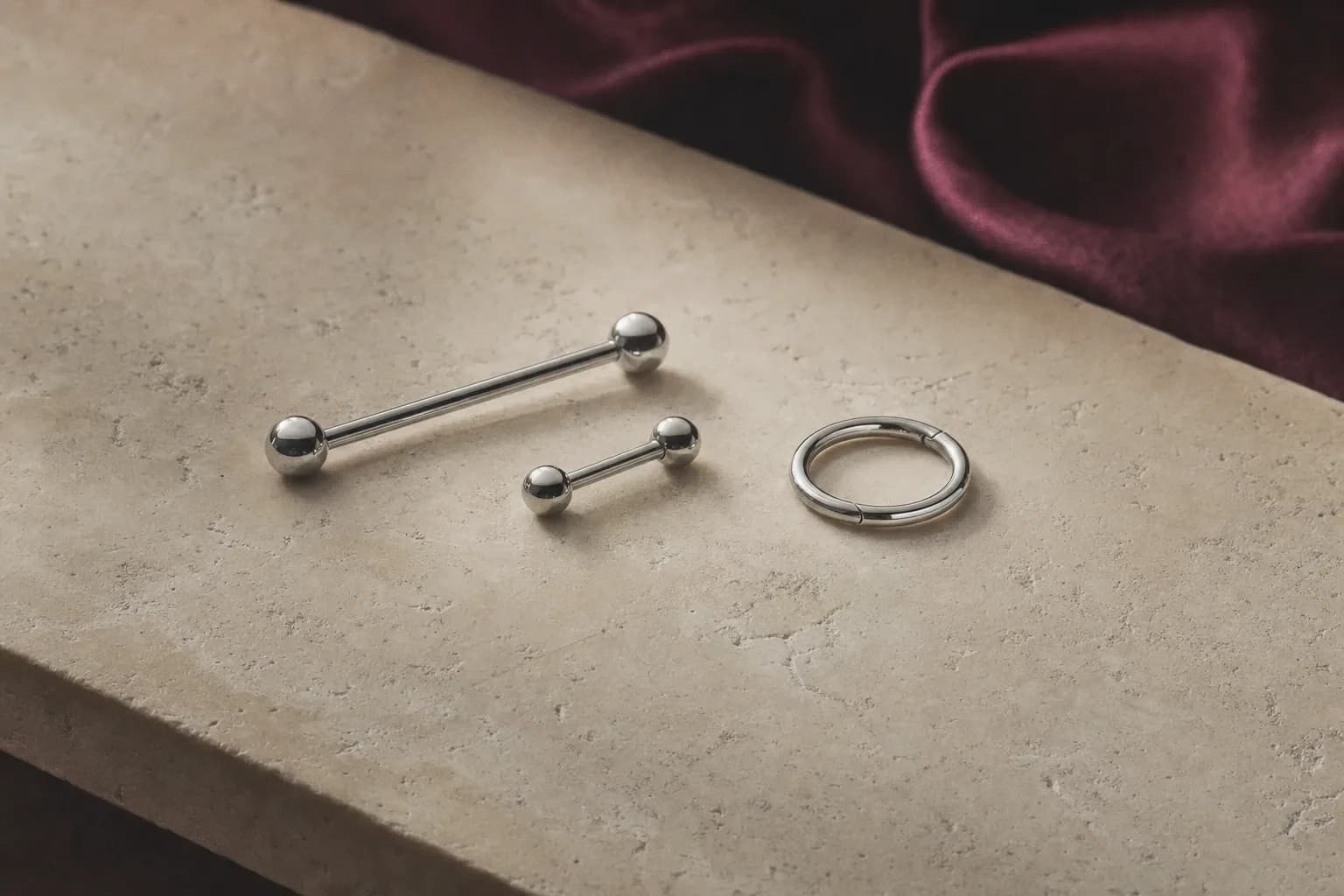 Zwei gerade silberfarbene Barbells und ein geschlossener Ring auf hellem Travertin, dahinter unscharfe pflaumenfarbene Seide